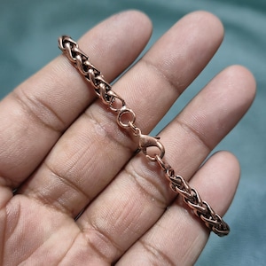 Puede incluir: Una pulsera de cadena de color cobre con un diseño tejido. La pulsera presenta un cierre de mosquetón. El metal tiene un tono marrón rojizo cálido. La pulsera se muestra sobre un fondo borroso.