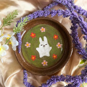 May include: A handmade ceramic plate with a painted bunny and flower design. The plate features a white bunny in the center, surrounded by red and pink flowers on a green background. The plate has a brown rim and is surrounded by purple flowers.