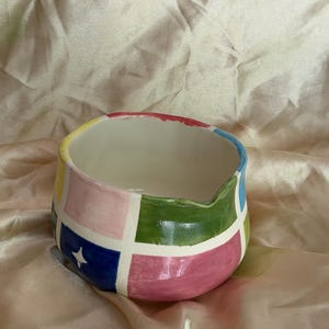 May include: A ceramic bowl featuring a hand-painted grid pattern with alternating squares of pink, green, blue, and yellow. The exterior surface includes a small white four-pointed star graphic on one of the blue squares and a small spout.