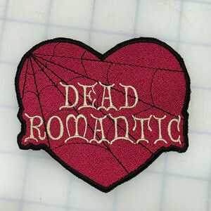 May include: A heart-shaped embroidered patch with a dark red background featuring a black spiderweb pattern. The text DEAD ROMANTIC is stitched in a cream-coloured, decorative font across the centre, all bordered by a thick black edge.