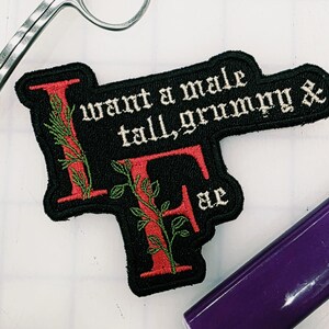 May include: A black embroidered patch featuring the red letters I and F intertwined with green vines. The text reads: I want a male tall, grumpy & Fae. The patch has a black border and is positioned near a metal tool and a purple object.
