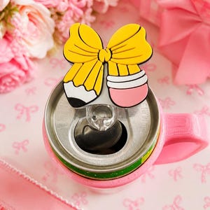 May include: A pink can cooler with a yellow bow-shaped pencil topper. The pencil has a black tip, white middle, and pink eraser. The background includes pink roses and a gift box, creating a cheerful, feminine aesthetic.
