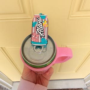 May include: A pink can cooler with a handle holds a beverage can. A decorative can-shaped topper with the word "Alani" in white script, pink stripes, and floral accents sits on top of the can.