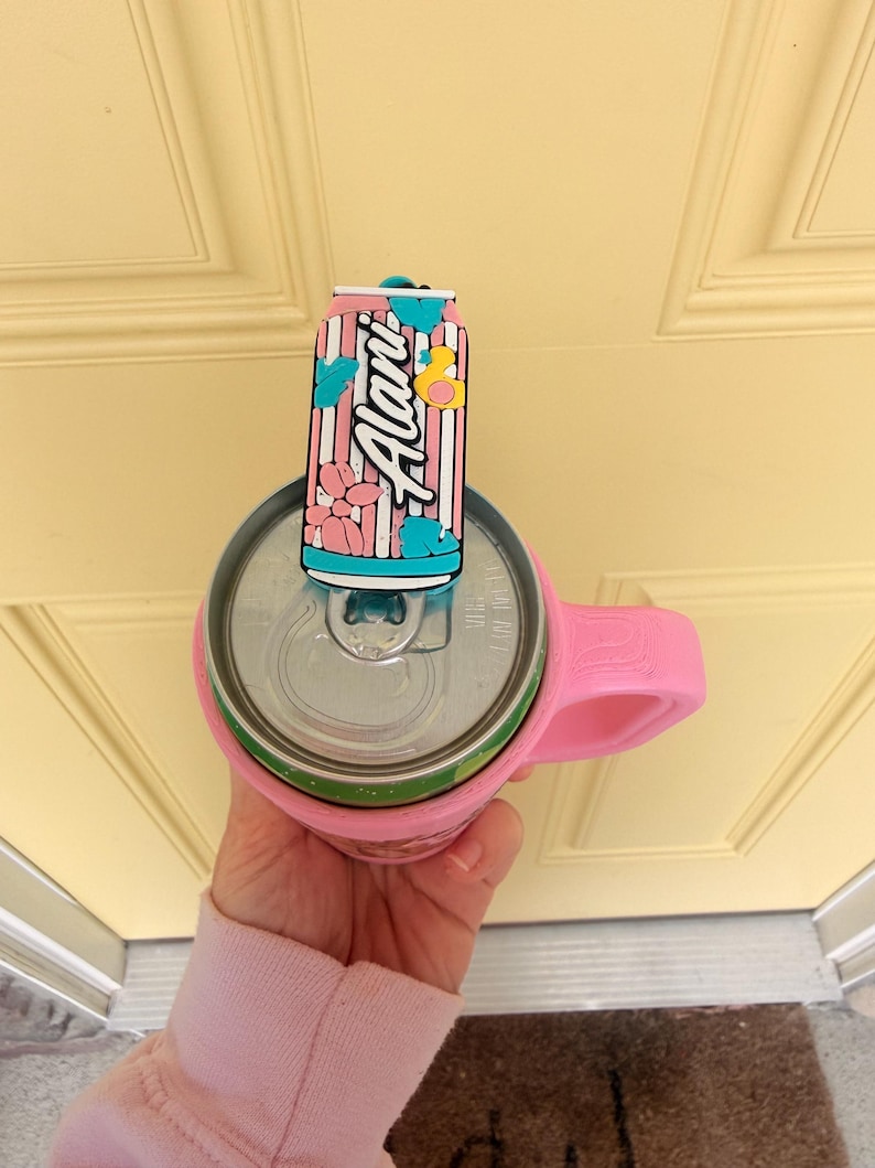 May include: A pink can cooler with a handle holds a beverage can. A decorative can-shaped topper with the word "Alani" in white script, pink stripes, and floral accents sits on top of the can.