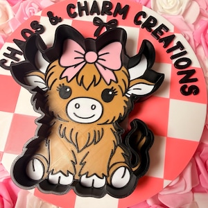 May include: A round, white and pink checkerboard sign with the text "Chaos & Charm Creations" in black. A brown and black Highland cow with a pink bow is in the center. The background is filled with pink and white roses.