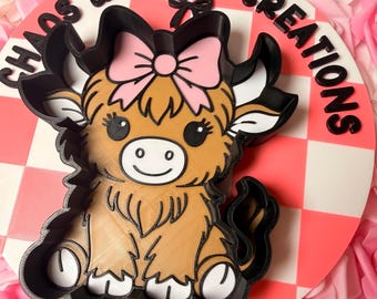 May include: A round, white and pink checkerboard sign with the text "Chaos & Charm Creations" in black. A brown and black Highland cow with a pink bow is in the center. The background is filled with pink and white roses.