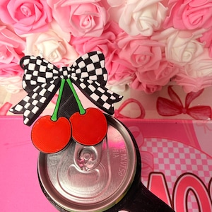 Puede incluir: Un lazo a cuadros blanco y negro con dos cerezas rojas encima de una lata de refresco. La lata es plateada con un asa negra. El fondo es un arreglo floral rosa y blanco.