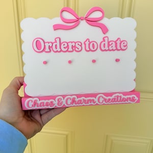 May include: A white rectangular sign with scalloped edges and a pink bow on top. The text "Orders to date" is in pink, with four small pink pegs below. The base is pink with the white text "Chaos & Charm Creations" displayed on the front.