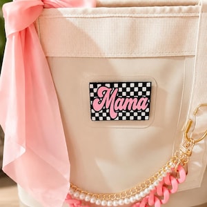 May include: A beige tote bag featuring a rectangular patch with a black and white checkerboard pattern and the word Mama in pink script. A light pink scarf is tied to the top, and a decorative chain made of gold links, white pearls, and pink links.
