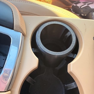 Könnte beinhalten: Ein Getränkehalter im Auto mit schwarzem Innenraum und einem grau strukturierten Einsatz. Der Getränkehalter befindet sich in einer beigefarbenen Konsole, wobei links eine silberne Zierleiste zu sehen ist. Sonnenbrillen liegen auf dem perforierten Ledersitz im Hintergrund.