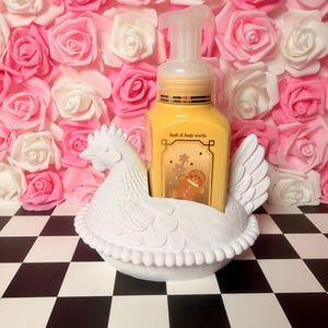 Puede incluir: Un jabonero blanco con forma de gallina que sostiene un dispensador de jab&oacute;n amarillo con el texto "bath & body works". El fondo presenta una pared de rosas rosas y blancas, y la base es un patr&oacute;n de tablero de ajedrez en blanco y negro.