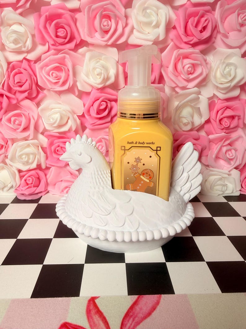 Puede incluir: Un jabonero blanco con forma de gallina que sostiene un dispensador de jab&oacute;n amarillo con el texto "bath & body works". El fondo presenta una pared de rosas rosas y blancas, y la base es un patr&oacute;n de tablero de ajedrez en blanco y negro.