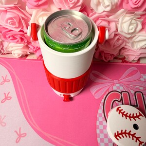 May include: A white and red drinks cooler containing a green and silver can. The cooler has red handles and a red base. A baseball-shaped coaster is on a pink surface with a bow design.