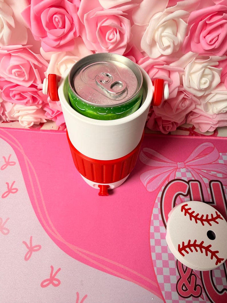 May include: A white and red drinks cooler containing a green and silver can. The cooler has red handles and a red base. A baseball-shaped coaster is on a pink surface with a bow design.