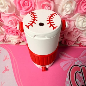 May include: A white and red baseball-themed tumbler. The top features red stitching details, and the body has a red band. The tumbler is set against a backdrop of pink and white roses.