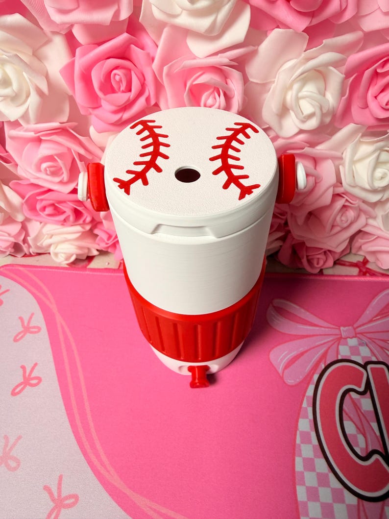 May include: A white and red baseball-themed tumbler. The top features red stitching details, and the body has a red band. The tumbler is set against a backdrop of pink and white roses.