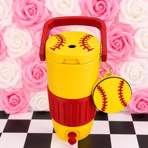 May include: A yellow softball water cooler with red handle and accents. The lid features red stitching details, and a matching softball charm hangs from the side. The cooler has a red base and a spigot for dispensing liquids.