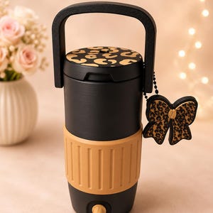 May include: Black and tan insulated beverage dispenser with a handle and a decorative bow charm. The lid and bow charm feature a gold leopard print. The dispenser has a tan band around the middle and a spigot at the bottom.