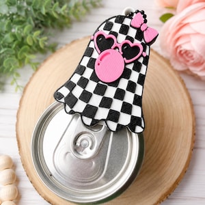 May include: A ghost-shaped beverage can opener topper featuring a black and white checkered pattern, pink heart-shaped sunglasses, a pink bubble gum accent, and a small pink bow. The accessory is designed to fit over the tab of a metal soda can.