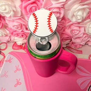 Puede incluir: Un enfriador de latas rosa con asa sostiene una lata de refresco, coronada con una tapa con temática de béisbol. La tapa de béisbol es blanca con costuras rojas. El enfriador se encuentra sobre una superficie rosa con un patrón de lazo, contra un fondo de rosas rosas y blancas.