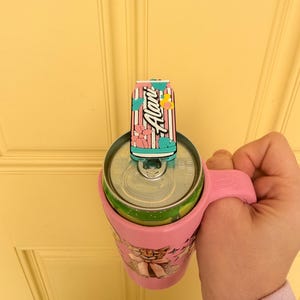 May include: A pink drink holder with a handle, holding a green and silver can. A decorative can-shaped topper is on top, with the word "Alani" in white script. The holder has a cartoon graphic.