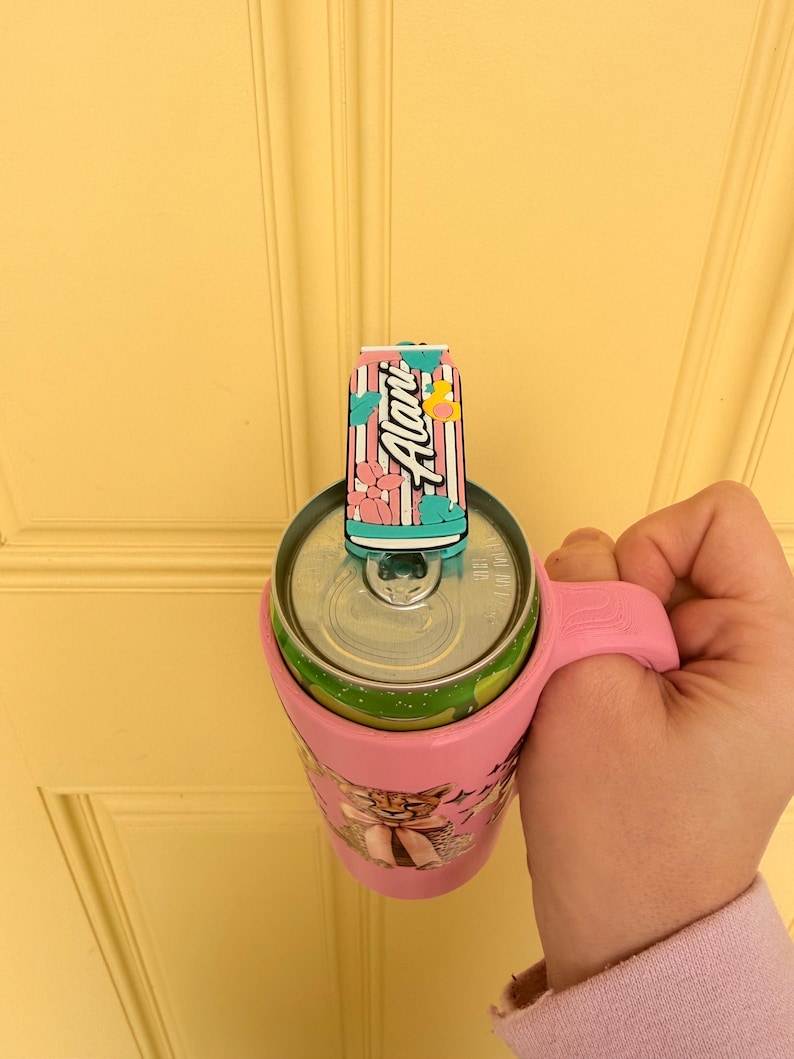 May include: A pink drink holder with a handle, holding a green and silver can. A decorative can-shaped topper is on top, with the word "Alani" in white script. The holder has a cartoon graphic.