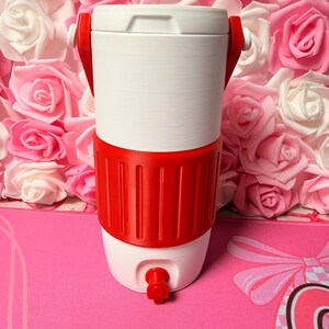 May include: A white and red water cooler with a tap. The cooler has a white top and bottom with a red band around the middle. Red handles are attached to the top. The cooler is set against a backdrop of pink and white roses.