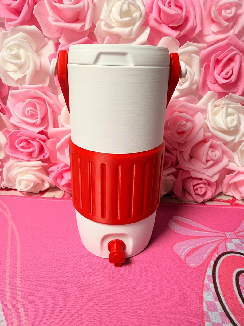 May include: A white and red water cooler with a tap. The cooler has a white top and bottom with a red band around the middle. Red handles are attached to the top. The cooler is set against a backdrop of pink and white roses.
