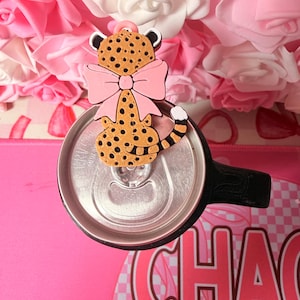 May include: A pink and brown cheetah-shaped charm with a pink bow, placed on top of a silver can. The charm has black spots and a white-tipped tail. The background features pink and white roses and the word "CHAOS".