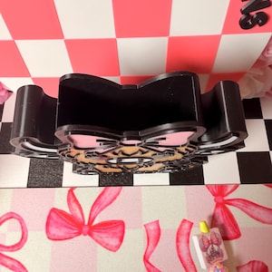 May include: Black desk organizer with a bow-shaped design and a pink and tan bow accent. The organizer is made of a sturdy material and has multiple compartments. The background features a pink and white checkered pattern.