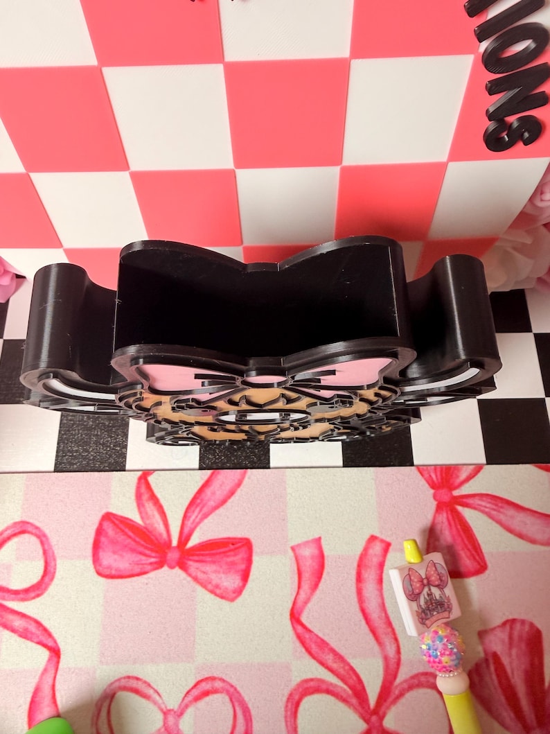 May include: Black desk organizer with a bow-shaped design and a pink and tan bow accent. The organizer is made of a sturdy material and has multiple compartments. The background features a pink and white checkered pattern.