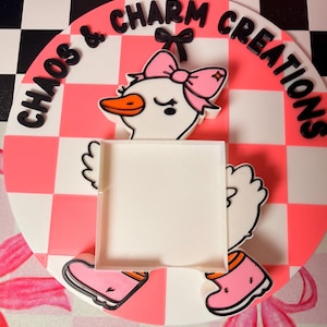May include: A round, white and pink sign with the text "CHAOS & CHARM CREATIONS" in black. The sign features a cartoon goose with a pink bow, boots, and a blank square. The background is a pink and white checkerboard pattern.