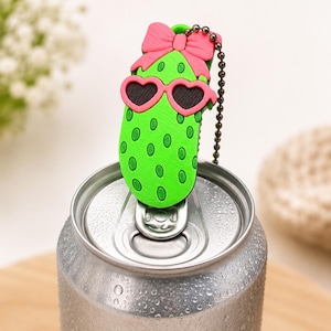 May include: A novelty can topper with a green pickle design, pink heart-shaped sunglasses, and a bow. The pickle has green spots and is attached to a silver chain. The topper is on a silver can.