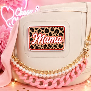 May include: A beige bag featuring a rectangular patch with a leopard print background and the word Mama in white script with a red outline. The bag is decorated with a layered strap consisting of a gold metal chain, a string of white pearls, and a thick pink plastic chain.