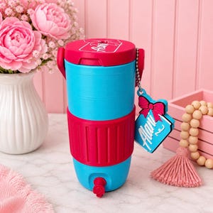 May include: A two-tone water cooler in blue and red, with a matching lid and base. The cooler has a spigot and a tag that says "Alani". The background includes pink flowers and a pink wall.
