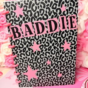 May include: A disc-bound notebook featuring a grey and black leopard print pattern on the cover. The word BADDIE is printed in black letters on a pink rectangular background. Several pink stars are scattered across the cover, and pink plastic discs secure the pages along the left side.