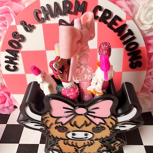 May include: A decorative display featuring a black and white checkered base, a pink and white checkered backdrop with the text "Chaos & Charm Creations", and a brown and tan Highland cow-shaped container filled with various pink and sparkly accessories.