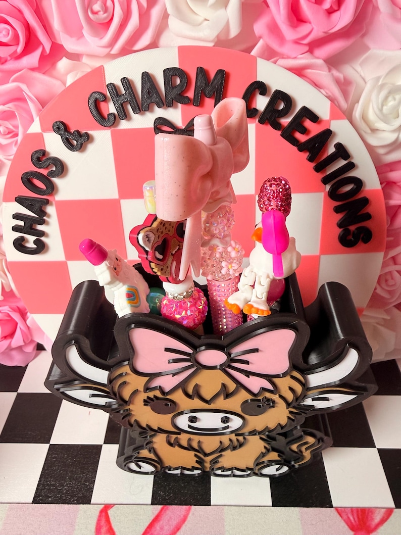 May include: A decorative display featuring a black and white checkered base, a pink and white checkered backdrop with the text "Chaos & Charm Creations", and a brown and tan Highland cow-shaped container filled with various pink and sparkly accessories.