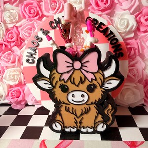 May include: A brown and black Highland cow-shaped container with a pink bow, holding small decorative items. The container sits in front of a pink and white checkered background with the words "Chaos & Creations". The background is a wall of pink and white roses.