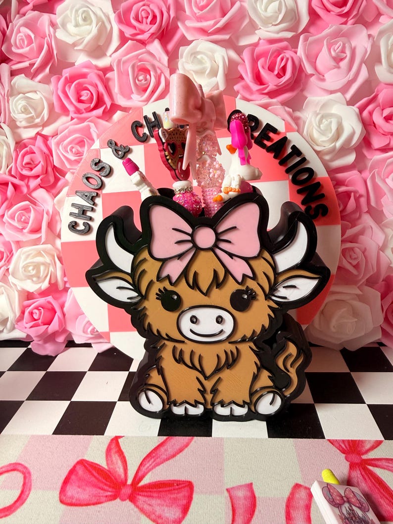 May include: A brown and black Highland cow-shaped container with a pink bow, holding small decorative items. The container sits in front of a pink and white checkered background with the words "Chaos & Creations". The background is a wall of pink and white roses.