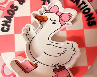 May include: A white cartoon goose wearing a pink bow and boots, set against a pink and white checkered background. The words "Chaos & Charm Creations" are in black letters around the edge of the circle.