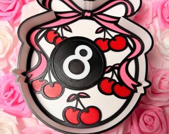 May include: A circular tray with a raised white border and a black outline. The center features a black billiard ball with a white number eight, surrounded by red cherries with green stems and pink ribbons tied in a bow at the top.