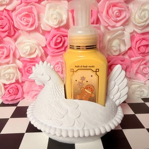 May include: A white ceramic chicken soap dispenser holder with a yellow soap bottle featuring a gingerbread man design. The background is a wall of pink and white roses, set on a black and white checkered surface.