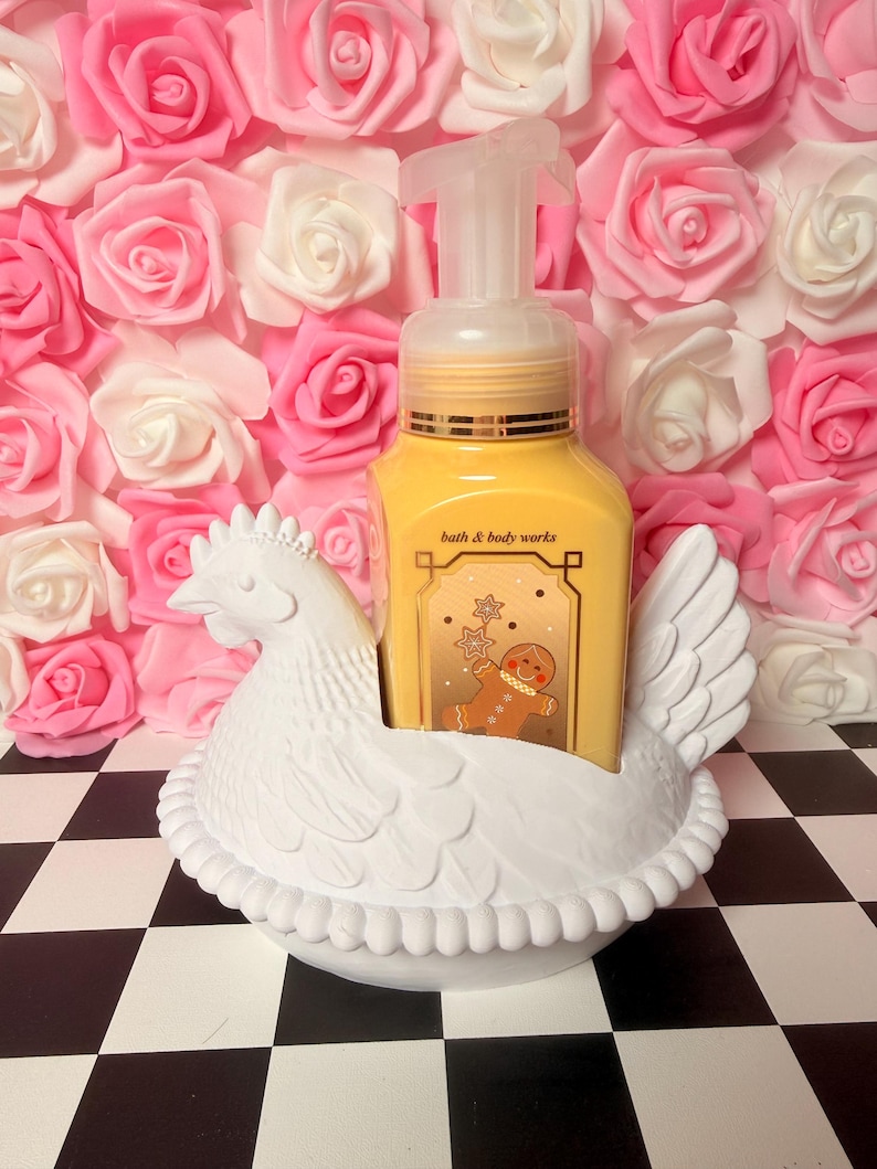 Puede incluir: Un soporte blanco de cer&aacute;mica con forma de gallina para dispensador de jab&oacute;n con una botella de jab&oacute;n amarilla con un dise&ntilde;o de hombre de jengibre. El fondo es una pared de rosas rosas y blancas, sobre una superficie a cuadros.