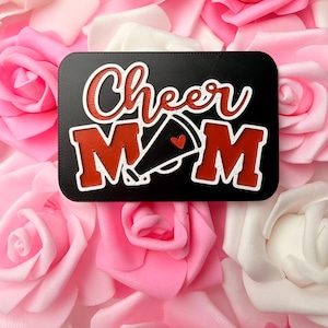 May include: A rectangular black sign featuring the white-outlined red text Cheer MOM, with a black megaphone icon replacing the letter O. The megaphone includes a small red heart detail. The sign is layered with a textured finish.