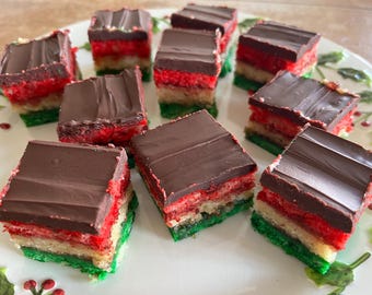 May include: A plate of colorful Italian rainbow cookies. Each square cookie has layers of green, white, and red cake, topped with a layer of chocolate. The cookies are arranged on a white plate with a holly design.