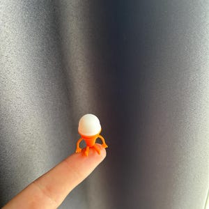 May include: A small, orange and white figurine resembling a creature with a rounded white head and orange body and legs. The figurine is perched on a fingertip against a gray background.