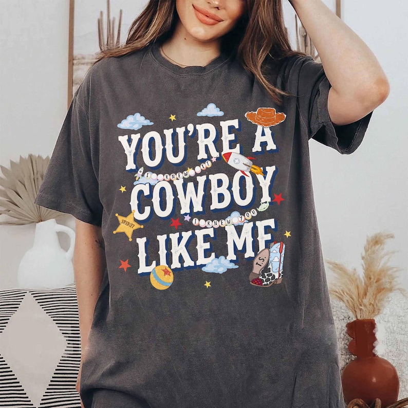 May include: A charcoal gray short-sleeve t-shirt featuring the white text "YOU'RE A COWBOY LIKE ME" surrounded by colorful graphic illustrations of a rocket, a cowboy hat, a sheriff star, a planet, and a patterned cowboy boot with clouds and stars.