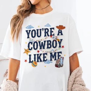 May include: A white short-sleeve t-shirt featuring the dark blue text "YOU'RE A COWBOY LIKE ME" with graphic elements including a brown cowboy hat, a rocket, a yellow sheriff star, a colorful ball, and a patterned cowboy boot with "T.S." on it.