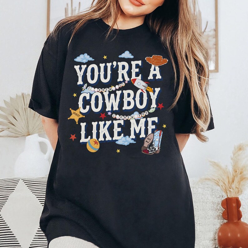 May include: A black t-shirt featuring the white text "YOU'RE A COWBOY LIKE ME" surrounded by colorful illustrations including a brown cowboy hat, a rocket ship, a yellow sheriff star, a blue and yellow ball, and a patterned cowboy boot.